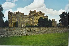 A large castellated stone house in front of which is a wall and an area of grass.