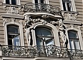 Farbige Nahaufnahme eines Fassadenteils mit drei Rundbogenfenstern und zwei nackten Frauenfiguren an den Seiten des mittleren Fensters. Unter allen Fenstern hängt ein Balkon und Ornamente sowie zwei Kopfreliefs befinden sich über den Fenstern.