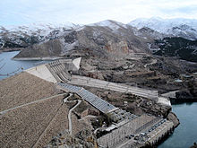 In the background brown mountains sprinkled with snow and a few clumps of dark green trees. Big pipes descend a thick concrete dam with electrical equipment at the base.