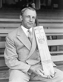 Cricketer Donald Bradman sits on a bench in a sports stadium and displays a cricket bat with his brand name on it. He wears a 1940s-style double-breasted suit with haircream. He is smiling and posing rather awkwardly.