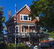 A three-story front-gabled ornate brick Queen Anne Style house seen from the front with two trees on either side casting shadows on it. Behind is a clear blue sky.