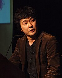 A man in his forties with black hair in a brown jacket addresses a seminar.