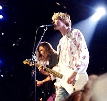 A side-view photograph of a blonde-haired man playing a guitar and singing into a microphone, along with a brown-haired man playing a bass guitar.