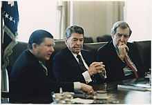 Three white men in suits sit at a table. All of them face right.