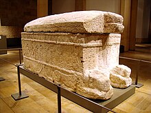 Three quarters view of an ornate stone sarcophagus. The carved relief shows a monarch sitting on a throne, his feet resting on a pedestal, an offerings table is depicted in front of the throne. The ruler's hand is raised in a greeting gesture; a procession of seven male persons bearing gifts faces the seated king. The sarcophagus sits on four lion effigies. The entire scene is framed by carved ropes and decorative details. The sarcophagus lid is convex and features a lion head and a line of Phoenician writing.