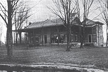 A house of wooden construction with a wrap-around front porch and trees in the front yard