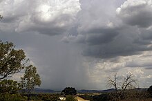 The sky is covered with dark clouds, from the middle of which a shaft of rain or hail comes down on hills on the horizon.