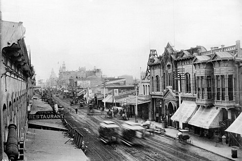Two horsecars pass in a blur c. 1889. Looking north along Main from just south of 1st Street. Grand Opera House at right. Towers of the United States Hotel at back, behind which the towers of the Baker Block.