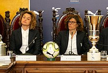 Two sitting women in a press conference with a ball and a trophy