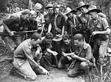 A group of soldiers kneel while another points to a mud model on the ground