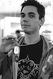 A black and white photo of a man with short dark hair, holding up military-style dog tags