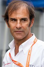 Emanuele Pirro wearing a white shirt with the first two buttons undone is looking down to his left with his eyes