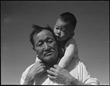 A black and white photo of an elderly, Japanese grandfather and his young Japanese grandson, on his shoulders at Manzanar Relocation Center, part of the photos that were impounded during the war