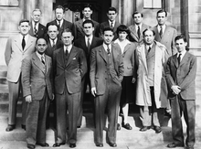 A group of people in suits standing in three rows on the steps in front of a stone building