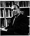 Ralph Ellison is pictured sitting in a chair before a bookcase. He is wearing a suit and has a mustache and receding hair-line.