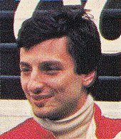 A man sporting black hair and wearing red racing overalls looking to the right of the camera