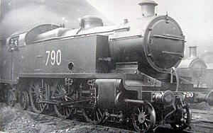 A side-and-front view of a large 2-6-4 steam locomotive at a locomotive depot. It is a tank locomotive with large rectangular water tanks either side of the boiler; they stretch from above the front driving wheels back to the cab. Members of the driving crew can be seen in the cab.