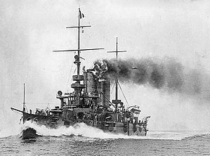 A small grey battleship traveling at full speed with smoke coming out of its two round funnels.