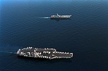 Aerial photograph of two aircraft carriers sailing in concert on calm water. The upper ship is smaller and has a small number of aircraft on its flight deck. The larger carrier, with a flat deck crowded with planes and helicopters, is towards the bottom.