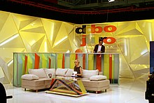 Croatian morning-show studio, with woman seated on sofa and man behind her standing at counter