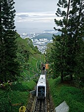 A funicular along a rail line flanked by trees at Penang Hill, with the cityscape visible in the background.