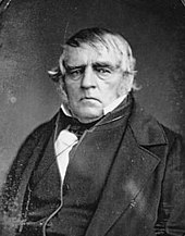Formal portrait showing the head and shoulders of a white-haired man wearing a dark cape or coat