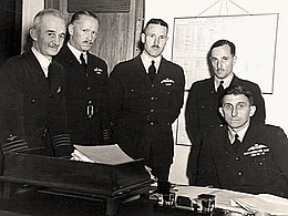 Five men in dark military uniforms behind a desk, four of whom are standing and one sitting