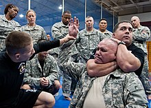 A man in fatigues rises his hand while another man in a black t-shirt holds his neck from behind with an arm to his neck and another to the back of his head. In the background men and women in fatigues observe.