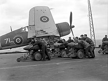 Black and white photograph of a group of men pushing bombs on trolleys on the deck of an aircraft carrier at sea. A single-engined aircraft is located immediately behind the men.