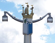 Post holding two lamps at the ends of two arms. The top of the post is decorated by a galleon; underneath the boat is a shield containing a white left and a blue right separated by a jagged line, on top of which rests a white bird carrying an olive branch.