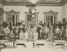 Photograph of 24 men both standing and seated surrounding a lone seated woman, in front of an American flag hanging between two Hawaiian flags