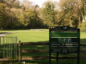 Cairn in the distance in sunshine, with trees in leaf to its left, right and rear. To its front lies flat ground of short grass. An asphalt path leads from the left past the tumulus. The shaded foreground has a kissing gate, a wooden fence and a Natural Resources Wales welcome sign in Welsh and English.