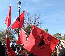 A crowd of people hold red, cloth flags above their heads at a rally. The signs read 'United Farm Workers. Si Se Puede' and display a stylized eagle symbol in the middle.