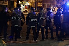 A group of police officers in helmets, carrying plastic shields with "POLICE" written on them, face the camera, on a city street at night. A bluish light glares into the right edge of the frame.