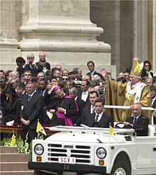 Farbige Momentaufnahme vom Papst in goldenem Papstgewand, der auf einem offenen Fahrzeug steht und in die Menge im Hintergrund winkt.