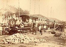 A black-and-white photo of a locomotive carrying and flanked by workers.
