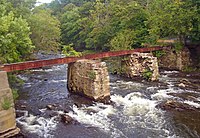 A rusted steel girder supported by two stone piers in the middle of a fast-flowing stream in a wooded area