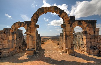 View through a triple-arched gate looking downhill to a street lined with ruins