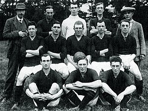 A faded photograph of a posed group of men, outdoors. At the front are three men seated on the ground, each dressed in sports clothing, a dark-coloured shirt, white shorts, dark socks and boots. In the middle row five men are sitting on benches, each dressed in sports clothing. Standing at the back are two men, weating hats and coats, and three men in sports clothing.
