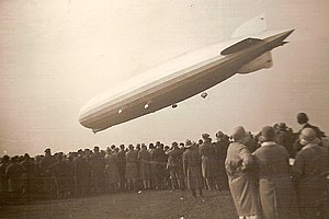 A black-and-white (sepia) image of a cigar-shaped airship flying from right to left over a crowd of people. Four of its five engines and the gondola are visible, as is the registration D-LZ127.