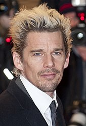 A Caucasian male with bleached hair, wearing a black overcoat. The collar of a gray suit is visible in the photo, with a white shirt and dark gray tie.