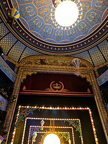 The interior of a grand theatre with a decorated roof, taken from the perspective of an audience member. You can see floating paper balloons above the seats that are a part of a festive performance.
