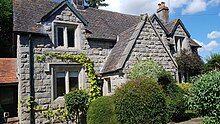 Frontale Farbfotografie von einem grauen Steinhaus. Das Haus mit seinen vier Dachgiebeln ist von Kletterpflanzen und Büschen umgeben.