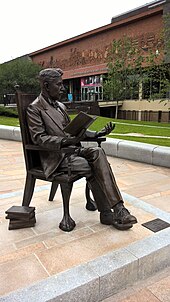 Statue of neatly-dressed, seated man holding an open book and gesturing, as if discussing it with someone