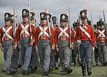 Photo shows marching men carrying muskets. They are dressed in red coats with white cross belts, gray trousers, and black shakos with brass frontplates.