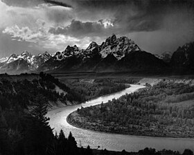 A black and white photograph shows a view over a river bend in foreground, to a rugged mountain range under cloudy skies