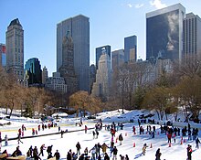 An outdoor skating rink with many people on the rink. There are skyscrapers in the background. This is the Wollman Rink in Central Park