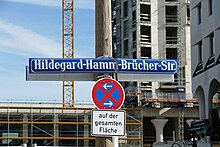 Foto eines blauen Straßenschildes mit weißer Schrift "Hildegard-Hamm-Brücher-Str." Unter dem Straßenschild ist ein Halteverbotsschild mit Pfeilen in beide Richtungen, darunter ein weißes Schild mit Schrift in Schwarz "auf der gesamten Fläche". Hinter den Schildern sieht man unscharf einen Ausschnitt einer Großbaustelle