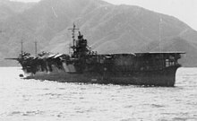 Photograph of a ship floating in the ocean taken from a location on the ocean surface some distance away. The upper deck of the ship is flat except for a superstructure visible near the center of the ship. The shoreline and some mountainous terrain are visible in the distance behind the ship; the sky is visible above the mountains.