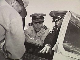 Three men in military uniforms with peaked caps crowding around the open cockpit of a military aircraft, two facing the camera and one with his back turned. The central figure in the portrait, facing the camera, has two rows of braid on the visor of his cap, and pilot's wings on the left breast pocket.
